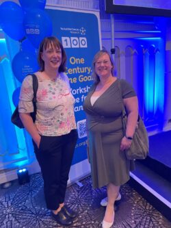 Hannah and Eva stand in front of a banner celebrating 100 years of Yorkshire Cancer Research at the Centenary Lunch